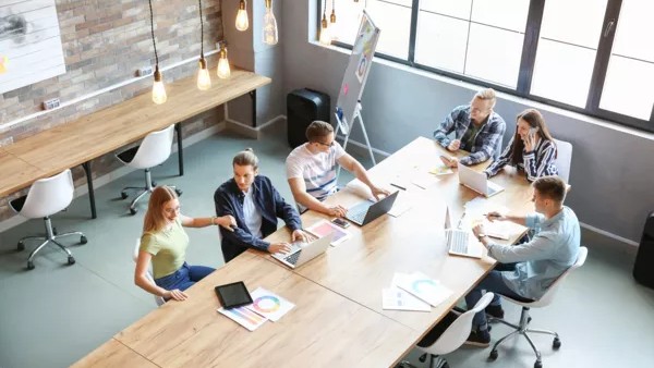 Symbolbild von einem Büro mit einem großen Schreibtisch, an dem sechs Personen sitzen und arbeiten.